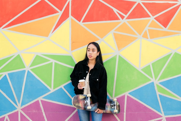 Joven mujer asiática con un café y una patineta en la mano apoyada en una pared colorida