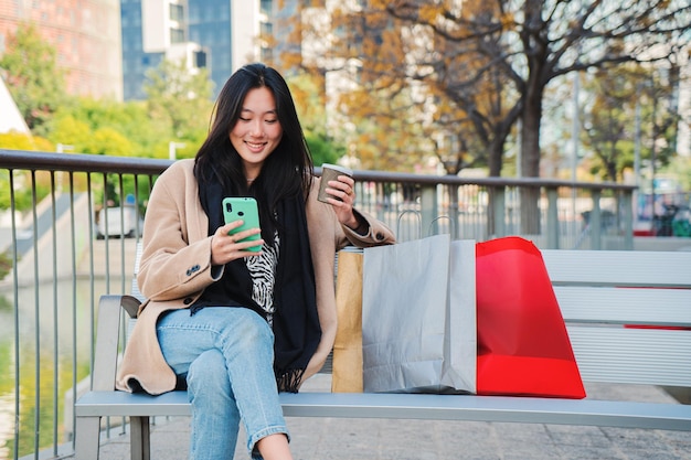 Joven mujer asiática despreocupada escribiendo un mensaje usando un teléfono celular y tomando un café después de comprar Adolescente enviando mensajes de texto con una aplicación de teléfono inteligente sentado en un banco al aire libre Concepto de comunicación