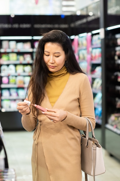 Joven mujer asiática elegante con bolso eligiendo un nuevo brillo de labios mientras sostiene el probador y lo va a aplicar en la mano en el supermercado de belleza