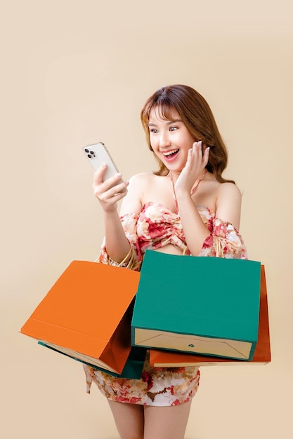 Joven mujer asiática enérgica que sostiene un teléfono inteligente de uso en blanco que busca una tienda minorista con coloridas bolsas de compras sobre fondo beige Concepto futurista de compras en línea