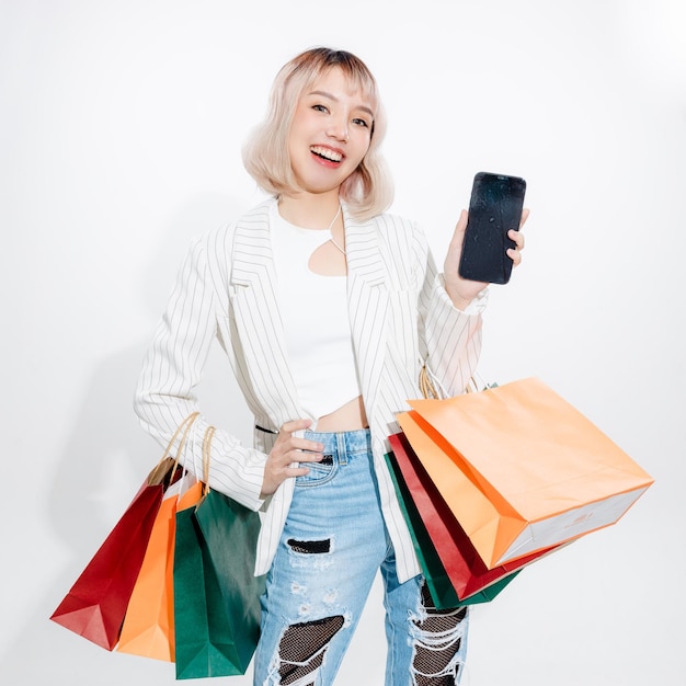 Joven mujer asiática enérgica que sostiene un teléfono inteligente de uso en blanco que busca una tienda minorista con coloridas bolsas de compras sobre fondo blanco Concepto futurista de compras en línea