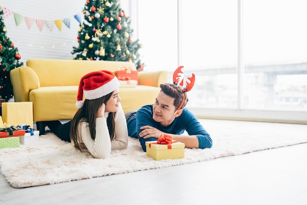 Joven mujer asiática feliz con un sombrero de Papá Noel con su novio con un regalo de Navidad mientras se acuesta en la alfombra con un árbol de Navidad en el fondo Foto con espacio para copiar