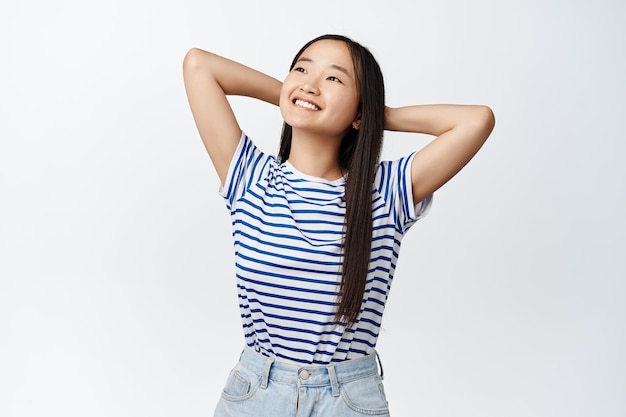 Joven mujer asiática feliz soñando, tendido con las manos detrás de la cabeza y descansando, mirando complacido en la esquina superior izquierda, sonriendo satisfecho en blanco