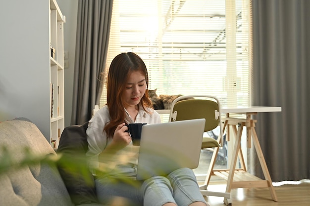 Joven mujer asiática independiente sentada en el sofá navegando por Internet revisando el correo electrónico en la computadora portátil por la mañana