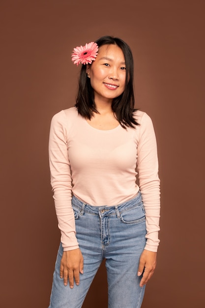 Joven mujer asiática en jeans azul y suéter blanco manteniendo los brazos frente a sí misma mientras está de pie frente a la cámara contra el fondo marrón