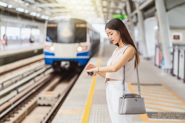 Joven mujer asiática pasajero escuchando música a través de un teléfono móvil inteligente y mirando el tren con reloj de mano en la estación de metro, estilo de vida japonés, chino, coreano, transporte y transporte