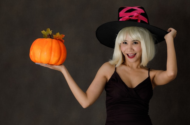 Joven mujer asiática con peluca rubia y sombrero de bruja con calabaza de Halloween riendo alegremente durante la fiesta sobre fondo negro