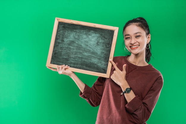 Joven mujer asiática presentando y apuntando a algo en una pizarra en blanco sobre una pared verde