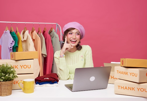 Joven mujer asiática puesta en marcha de pequeñas empresas independientes venta de ropa de moda con caja de paquetes y computadora portátil en la mesa y sentado aislado sobre fondo rosa Concepto de marketing y entrega en línea