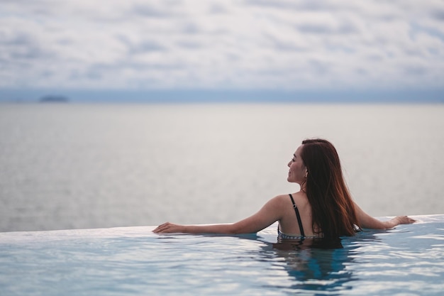 Una joven mujer asiática relajándose en la piscina infinita mirando una hermosa vista al mar