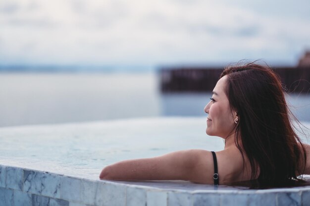 Una joven mujer asiática relajándose en la piscina infinita mirando una hermosa vista al mar