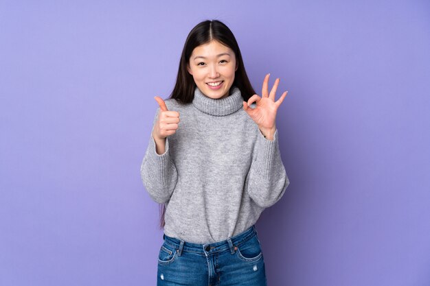 Joven mujer asiática sobre pared mostrando signo bien y pulgar arriba gesto