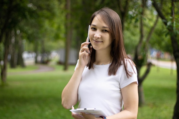 Joven mujer asiática sonriente hablando por teléfono móvil, de pie en una oficina moderna con un documento en la mano