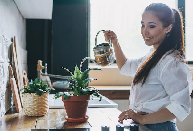 Joven mujer asiática sonriente con ropa informal regando plantas de interior en la cocina