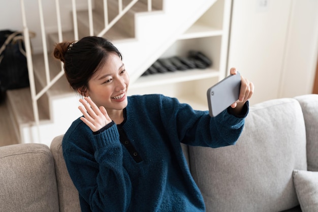 Joven mujer asiática sonriente sentada en un sofá acogedor sostiene la mano de la onda del teléfono celular saludando a la familia hace una videollamada hablando a través de una reunión virtual en casa Comunicación a distancia en el concepto de cuarentena