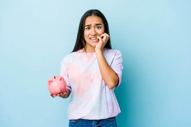 Joven mujer asiática sosteniendo un banco rosa sobre pared aislada mordiéndose las uñas, nerviosa y muy ansiosa