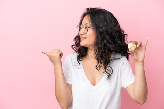 Joven mujer asiática sosteniendo un Bitcoin aislado sobre fondo rosa apuntando hacia el lado para presentar un producto