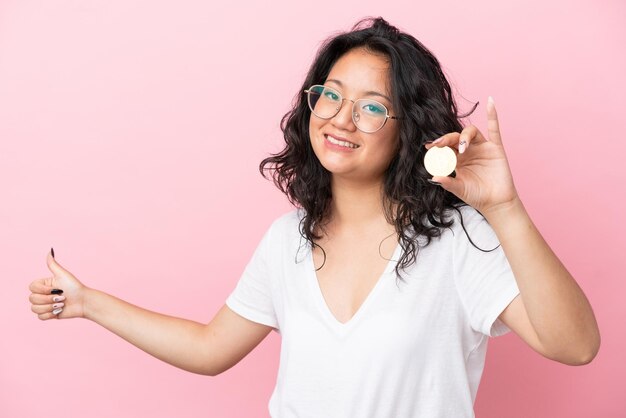 Joven mujer asiática sosteniendo un Bitcoin aislado sobre fondo rosa dando un pulgar hacia arriba gesto