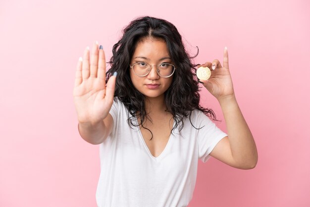 Joven mujer asiática sosteniendo un Bitcoin aislado sobre fondo rosa haciendo gesto de parada