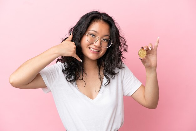 Joven mujer asiática sosteniendo un Bitcoin aislado sobre fondo rosa haciendo gesto de teléfono. Llámame señal