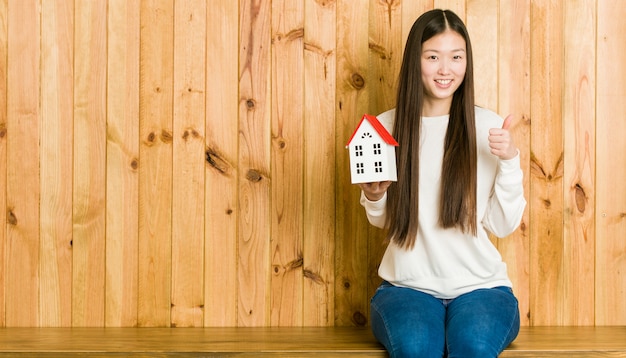 Joven mujer asiática sosteniendo un icono de la casa sonriendo y levantando el pulgar