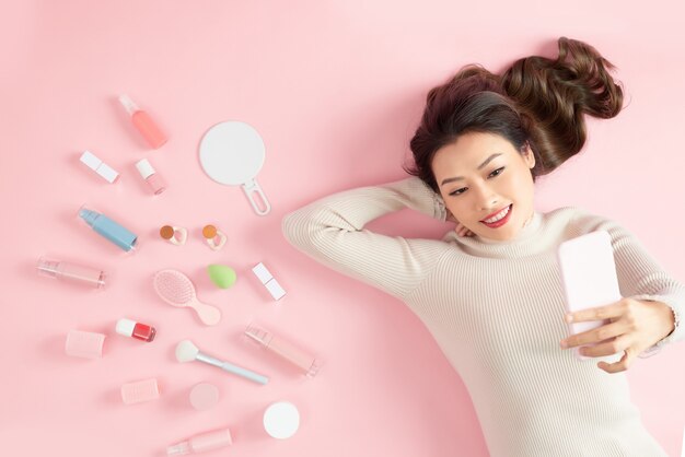 Joven mujer asiática tomando selfie con su teléfono y acostado en rosa con sus cosméticos