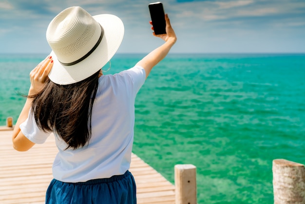 Joven mujer asiática usar sombrero de paja en estilo casual uso smartphone tomando selfie en muelle de madera. Vacaciones de verano en la playa del paraíso tropical. Chica feliz viaje de vacaciones.