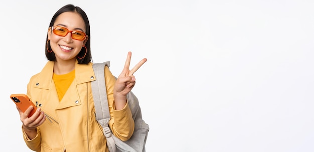 Joven mujer asiática viajera turista con gafas de sol sosteniendo mochila y teléfono móvil usando la aplicación en el teléfono inteligente de pie sobre fondo blanco