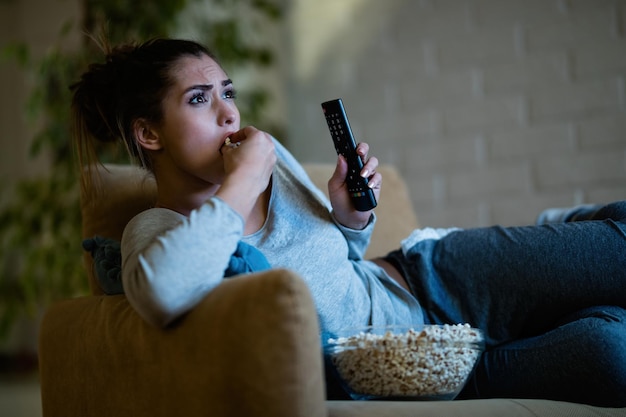 Joven mujer asustada viendo televisión y comiendo palomitas de maíz por la noche en casa