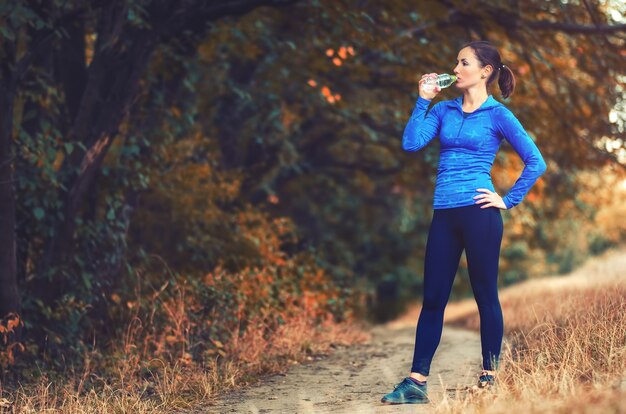 Una joven mujer atlética con una chaqueta deportiva azul con capucha y leggins negros bebe agua de la botella después de trotar