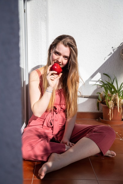 Joven mujer atractiva alegre en vestido casual de verano sentado en el balcón y comer sabrosa jugosa manzana roja