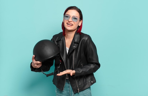 Joven mujer atractiva de cabello rojo sonriendo alegremente, sintiéndose feliz y mostrando un concepto en el espacio de la copia con la palma de la mano. concepto de motociclista