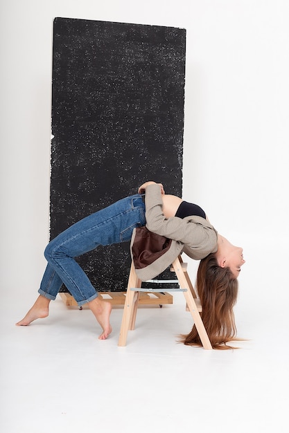 Joven mujer atractiva caucásica con cabello largo castaño en jeans, chaqueta de traje doblando su cuerpo sobre fondo blanco. Mujer flaca acostada en posición de puente sobre la escalera de madera en el estudio con los pies descalzos