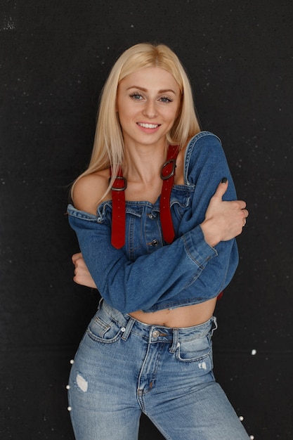 Joven mujer atractiva feliz con una sonrisa y pecas en ropa de jeans con jeans de moda cerca de la pared negra