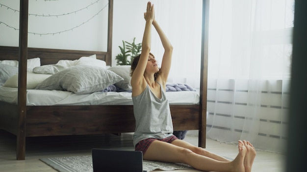 Joven mujer atractiva haciendo ejercicio de yoga y viendo una lección de tutorial en una computadora portátil en casa