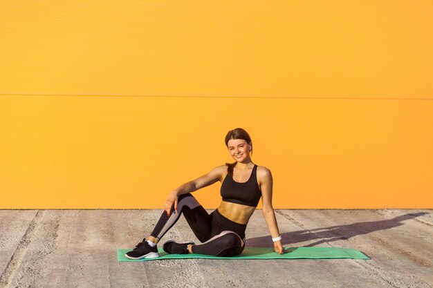 Joven mujer atractiva con ropa deportiva negra practicando ejercicios deportivos por la mañana en la calle, sentada en bandas verdes y descansando después del entrenamiento, mirando la cámara. fondo de pared naranja, exterior