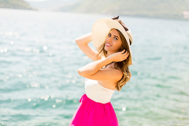 Joven mujer atractiva con un sombrero posa junto al mar en un día soleado