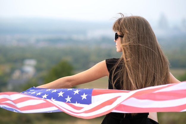 Joven mujer bastante americana con el pelo largo sosteniendo ondeando en el viento la bandera de EE.UU. en sus hombros descansando al aire libre disfrutando del cálido día de verano
