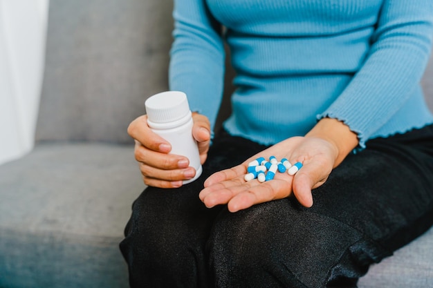 Joven mujer bastante asiática sosteniendo pastillas de medicina cuando se sienta en el sofá