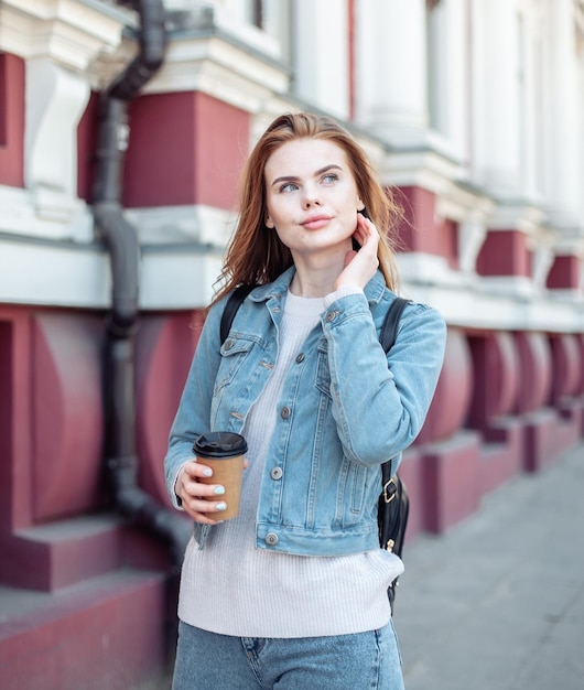 Joven mujer bastante atractiva con chaqueta vaquera sosteniendo una taza de café en la ciudad