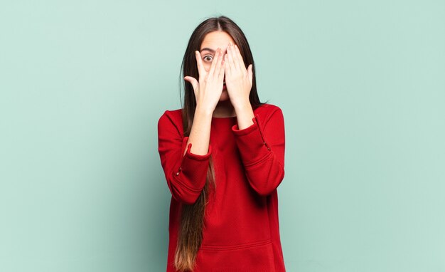 Joven mujer bastante casual que cubre la cara con las manos, mirando entre los dedos con expresión de sorpresa y mirando hacia un lado