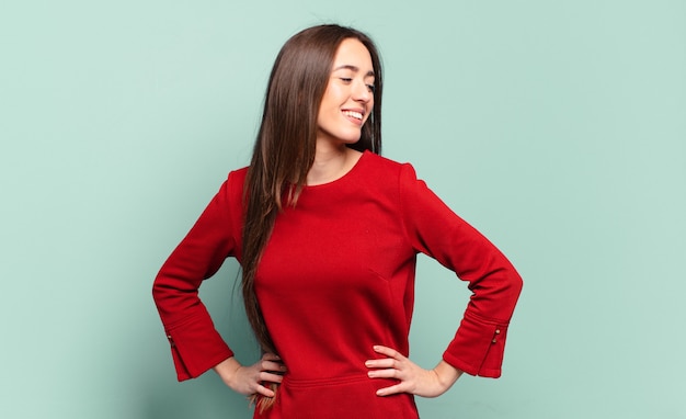 Joven mujer bastante casual que parece feliz, alegre y confiada, sonriendo con orgullo y mirando al lado con ambas manos en las caderas