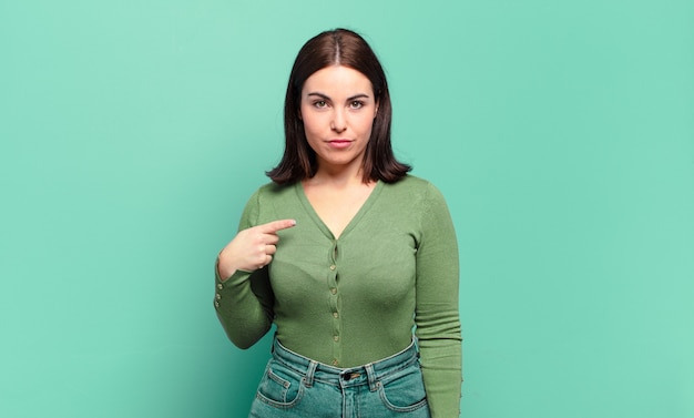 Joven mujer bastante casual que parece orgullosa, segura y feliz, sonriendo y apuntando a sí misma o haciendo la señal número uno