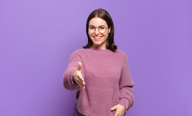 Joven mujer bastante casual sonriendo, luciendo feliz, confiada y amigable, ofreciendo un apretón de manos para cerrar un trato, cooperando