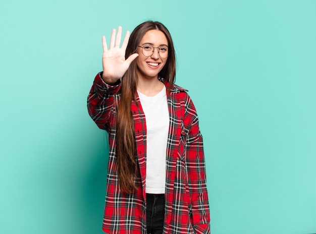 Joven mujer bastante casual sonriendo y mirando amistosamente, mostrando el número cinco o quinto con la mano hacia adelante, contando hacia atrás