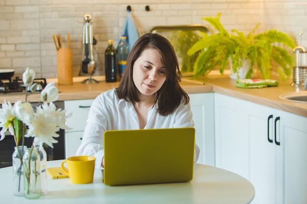 Joven mujer bastante caucásica hablando por teléfono sentada frente a la computadora portátil en la cocina. ama de casa de negocios. bebiendo café de una taza amarilla