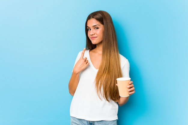 Joven mujer bastante caucásica sosteniendo un café para llevar apuntando con el dedo a usted como si invitara a acercarse.