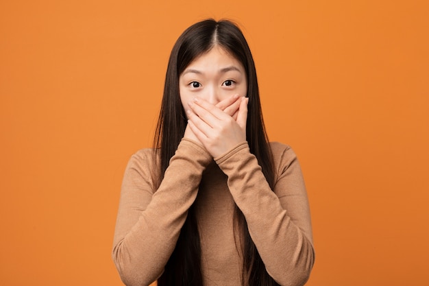 Joven mujer bastante china sorprendida cubriendo la boca con las manos.