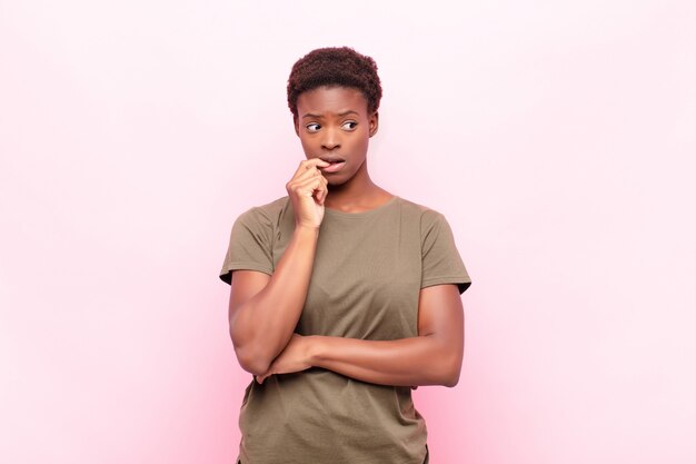 Joven mujer bastante negra con mirada sorprendida, nerviosa, preocupada o asustada, mirando a un lado hacia el espacio de la copia contra la pared rosa