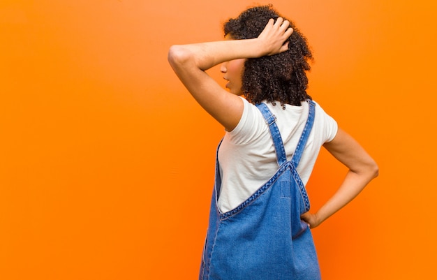 Joven mujer bastante negra pensando o dudando, rascándose la cabeza, sintiéndose perplejo y confundido, vista posterior o posterior contra la pared naranja